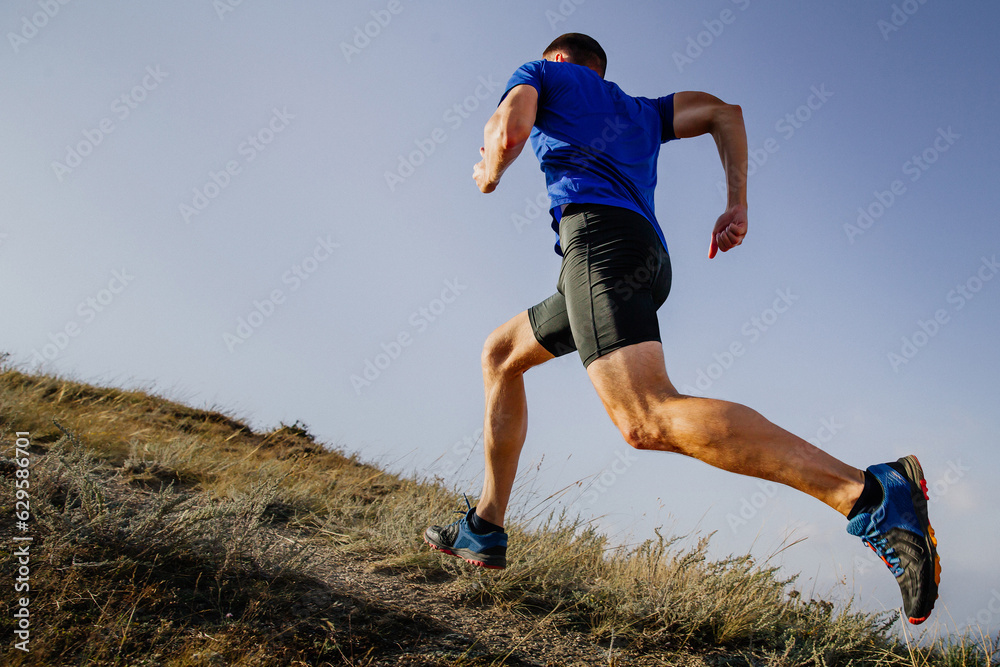 athlete runner running mountain uphill in blue shirt and black tights, background of sky