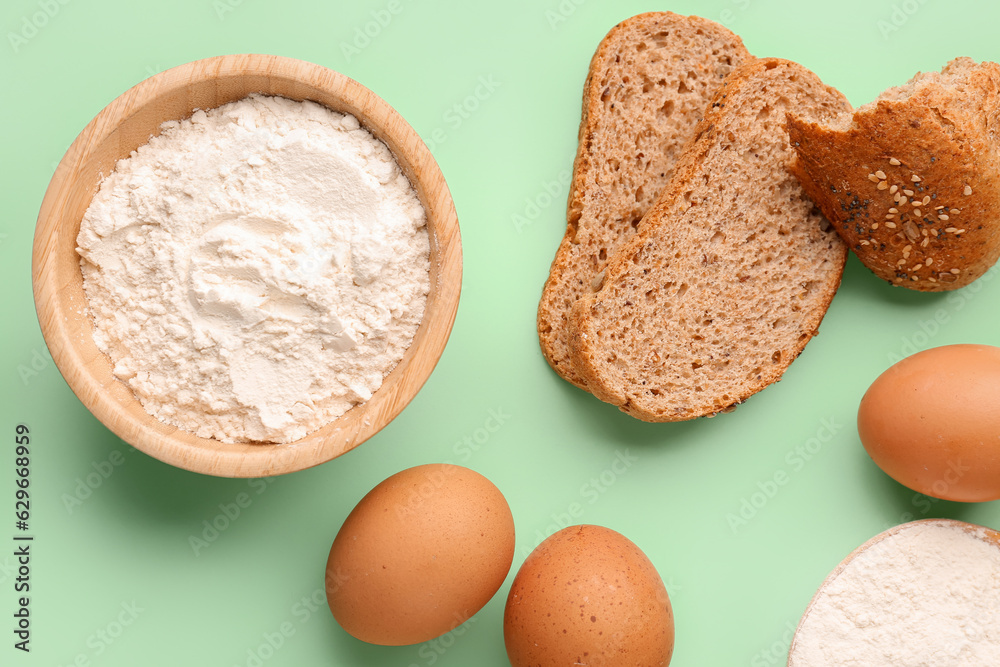 Wooden bowl with wheat flour, bread and eggs on green background