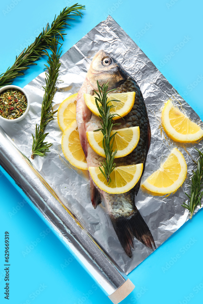 Aluminium foil roll with fresh raw fish, lemon slices, rosemary and spices on color background, clos