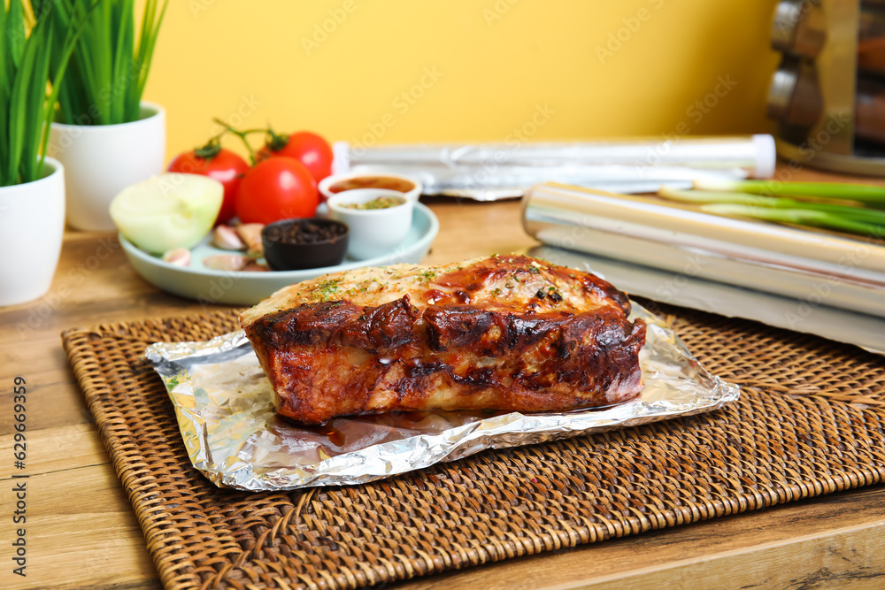 Aluminium foil with tasty baked meat, vegetables and spices on wooden table in kitchen