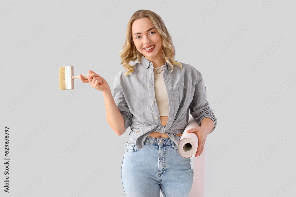 Young woman with wallpaper roll and brush on grey background