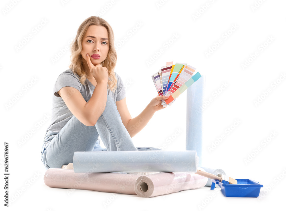 Upset young woman with color palettes and wallpaper rolls sitting on white background