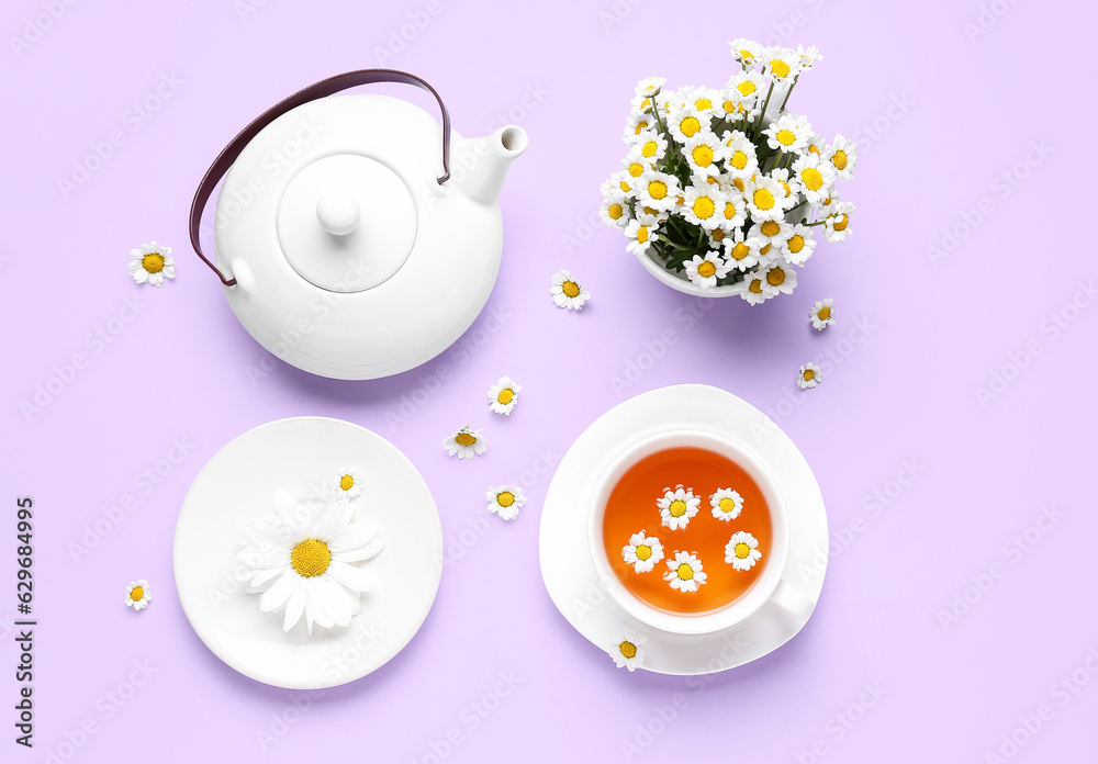 Teapot with cup of natural chamomile tea and flowers on lilac background