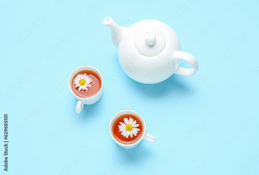 Teapot with cups of natural chamomile tea and flowers on blue background