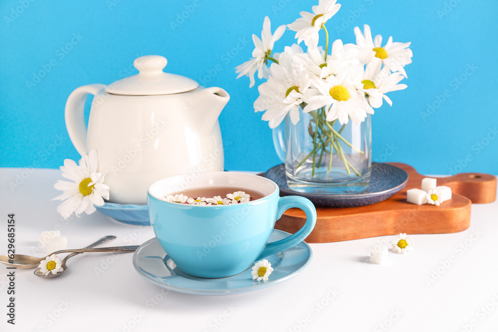 Wooden board with teapot, cup of natural chamomile tea and flowers on white table near blue wall