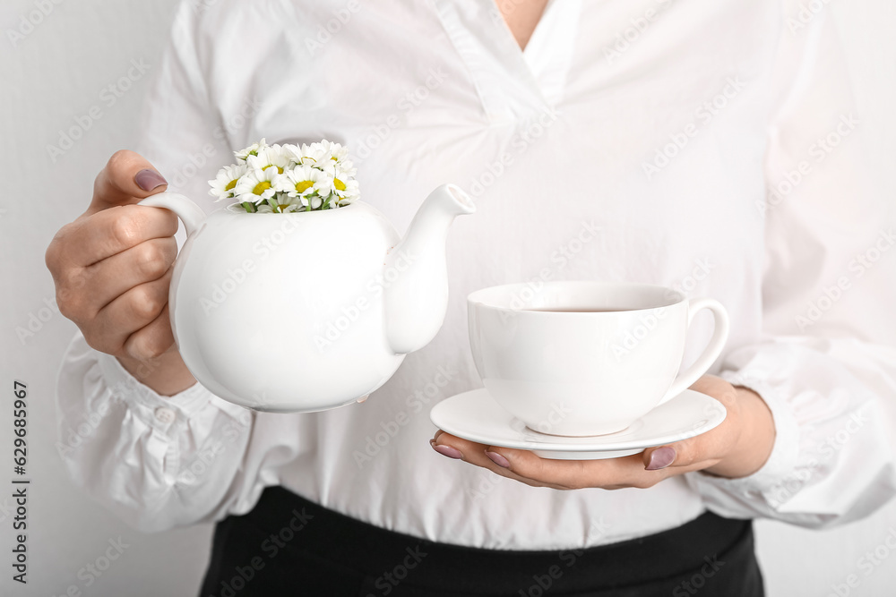 Beautiful young woman with teapot of chamomile tea and cup near white wall