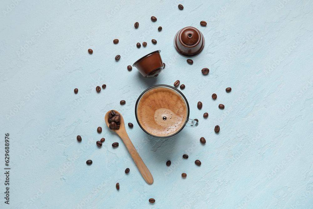 Cup of delicious coffee, wooden spoon, beans and capsules on blue table