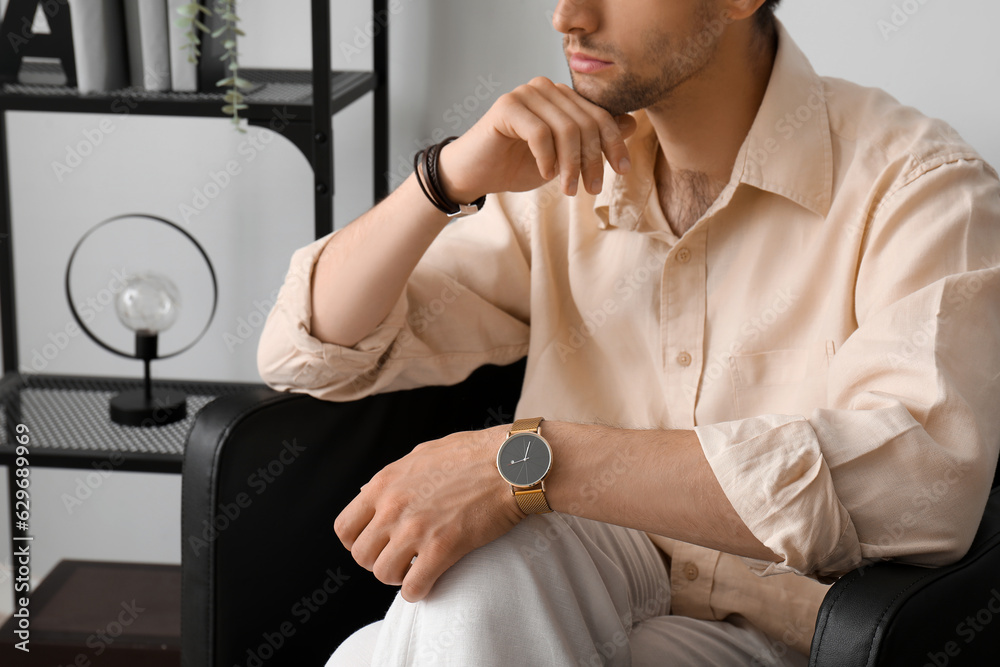 Stylish young man with wristwatch sitting on armchair in room