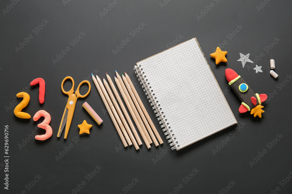 Blank notebook with rocket made of plasticine and different stationery on black chalkboard