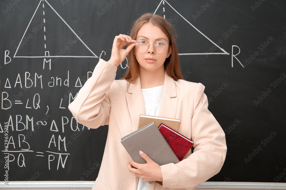 Young math teacher with books near blackboard in classroom