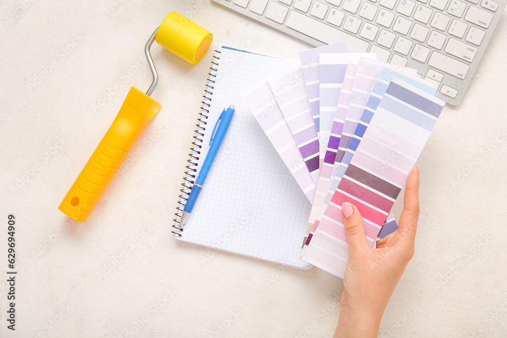 Hand with color palettes, PC keyboard, paint roller and notebook on light background