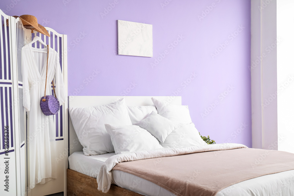 Interior of stylish bedroom with comfortable bed, white pillows and dress on folding screen