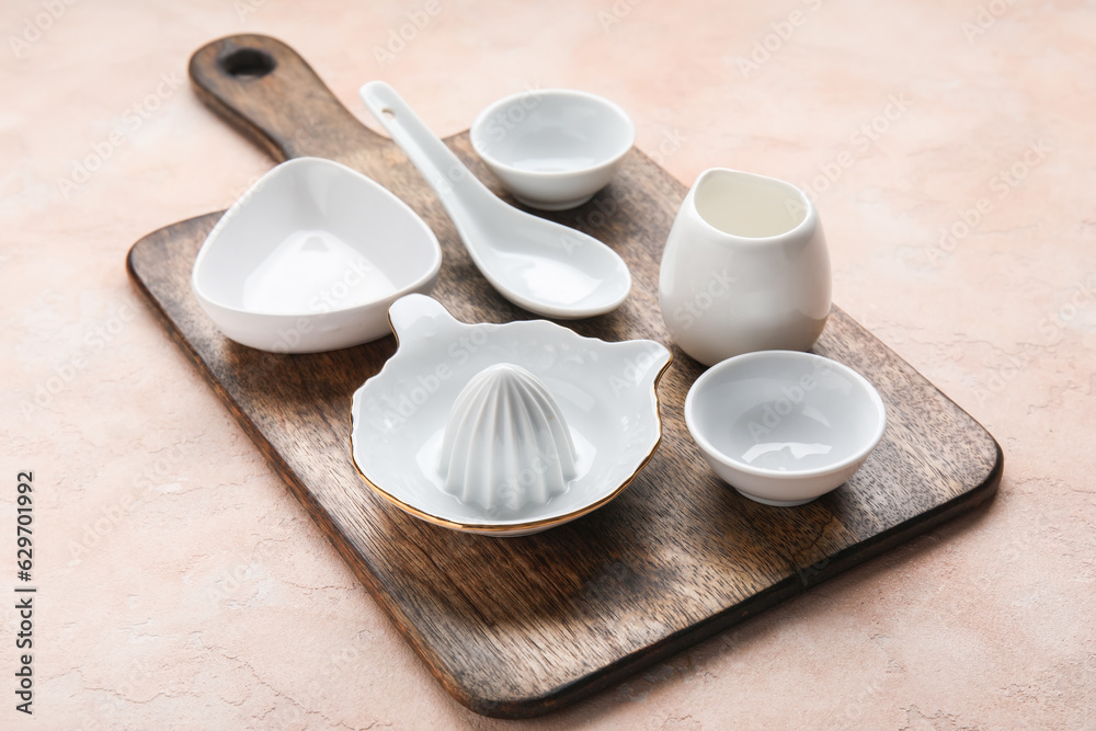 Wooden board with ceramic kitchen utensils on color background