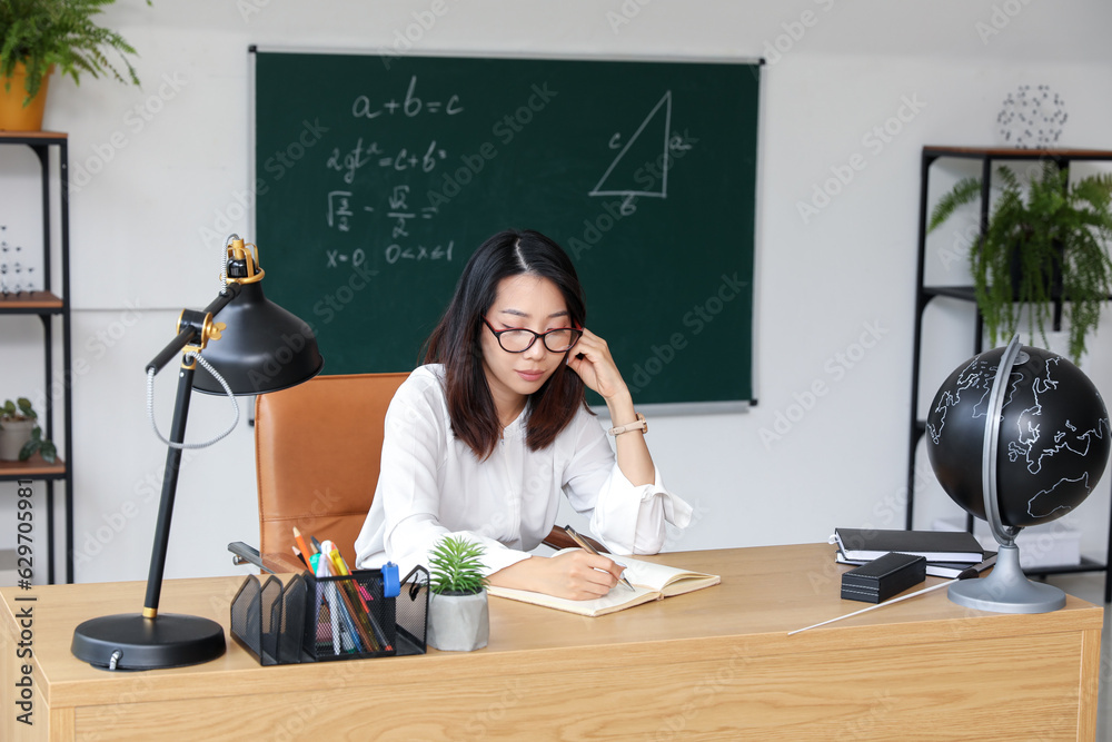 Female Asian teacher writing at table in classroom