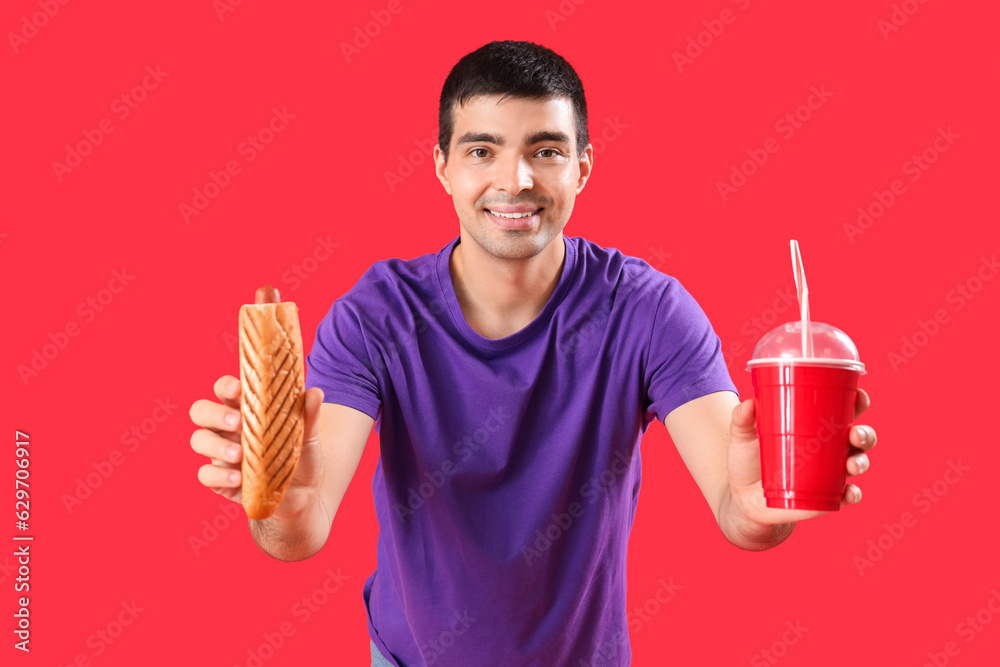 Happy young man with tasty french hot dog and drink on red background