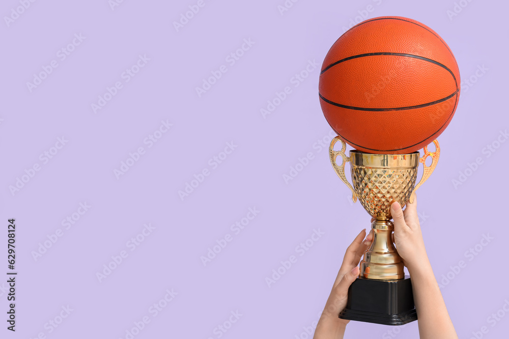 Female hands with gold cup and basketball on lilac background