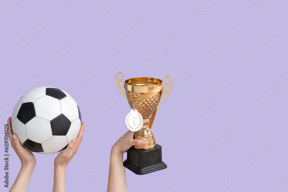 Female hands with gold cup, medal and soccer ball on lilac background