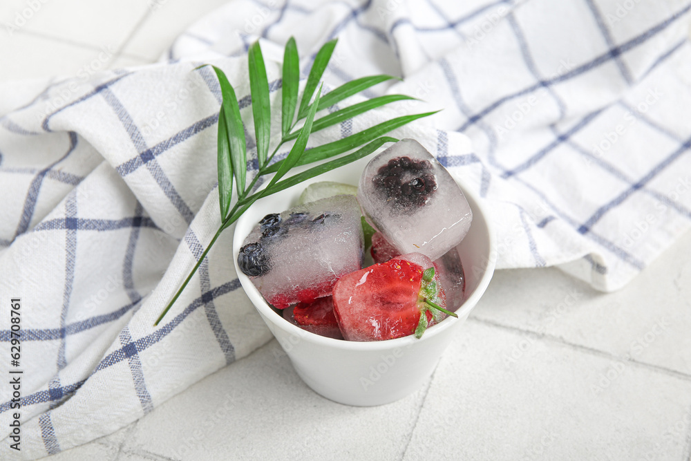 Bowl with frozen berries in ice cubes and palm leaf on white tile background