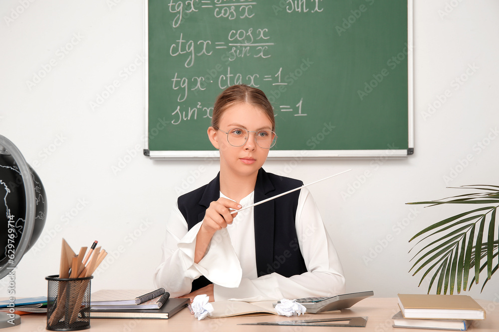 Young math teacher with pointer sitting at wooden table in classroom