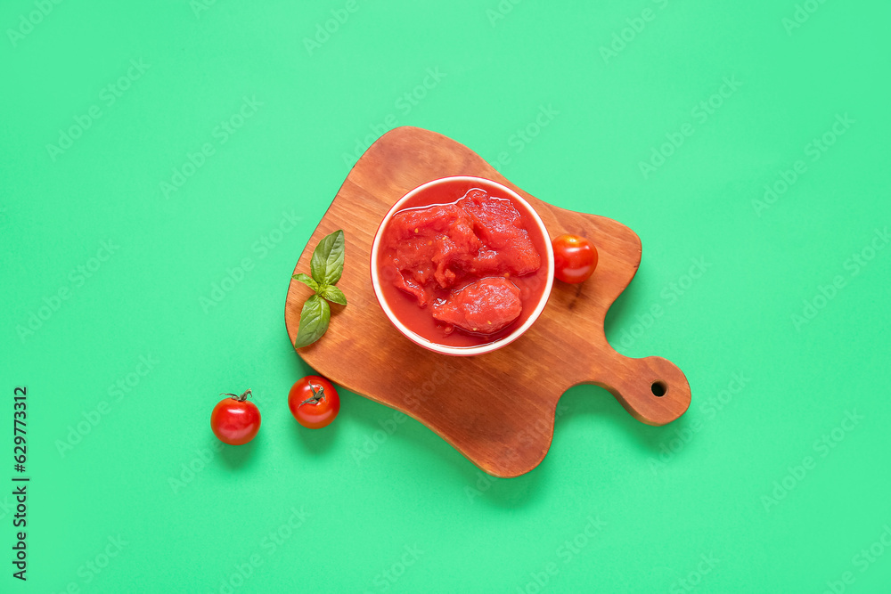 Wooden board with bowl of canned tomatoes on green background