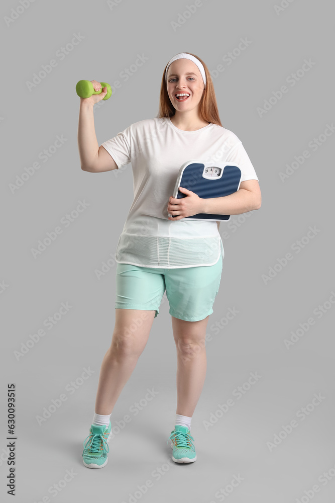 Young overweight woman with scales and dumbbell on grey background