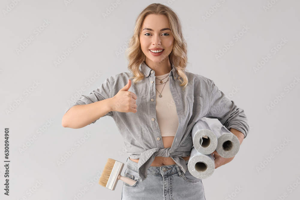 Young woman with wallpaper rolls showing thumb-up on grey background