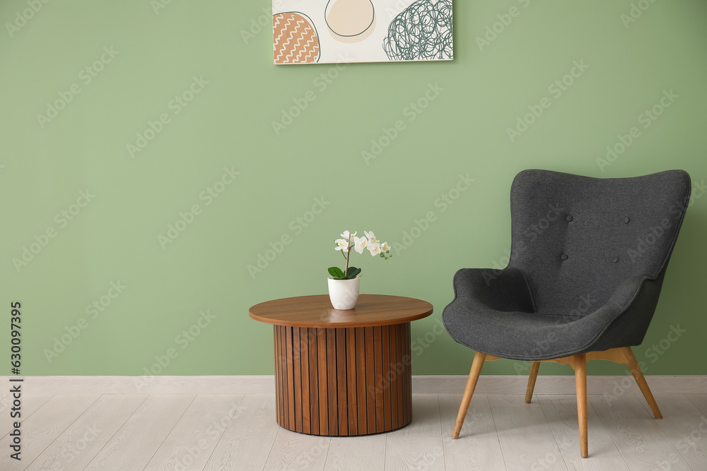 Dark grey armchair with wooden coffee table, orchid and painting near green wall