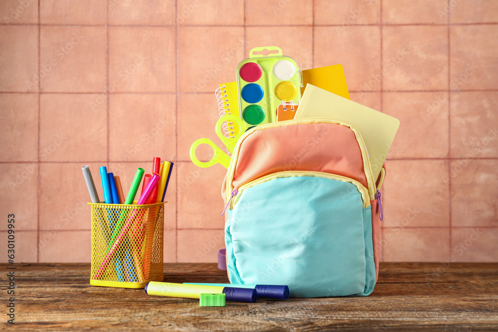 Colorful school backpack with notebooks, watercolors and cup of markers on wooden table near beige t