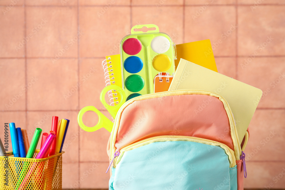 Colorful school backpack with notebooks, watercolors and cup of markers on beige tile background