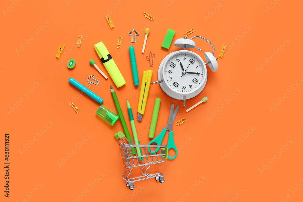 Alarm clock and shopping cart with different stationery on orange background