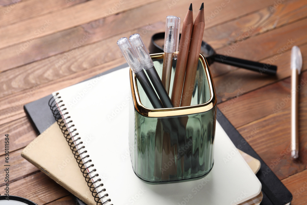 Glass holder with pens, pencils and notebooks on wooden table