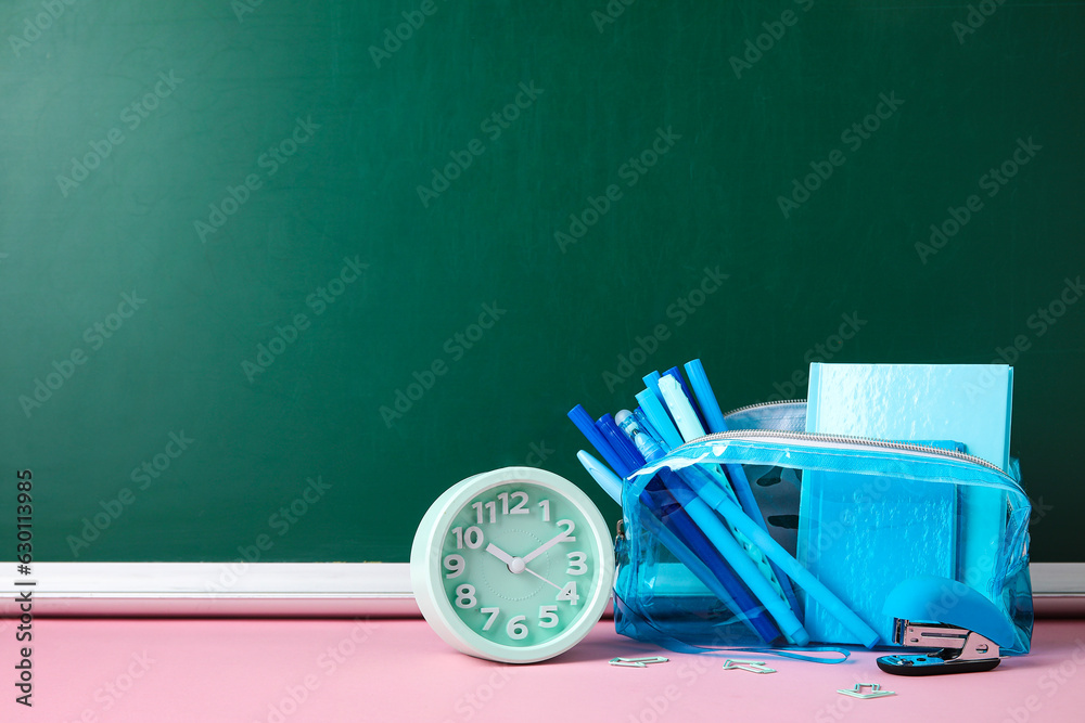 Pencil case with different school stationery and alarm clock on pink table near blackboard