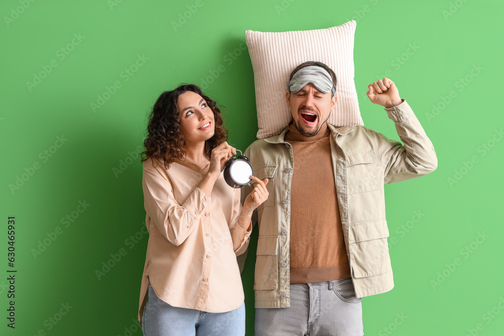 Young woman with alarm clock waking up her husband on green background