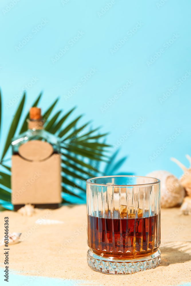 Glass of cold rum on sand against blue background