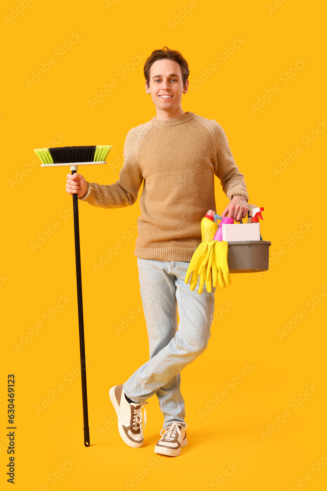 Young man with broom and bucket of cleaning supplies on yellow background