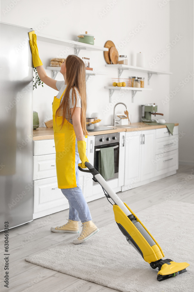 Young woman with vacuum cleaner wiping fridge in kitchen