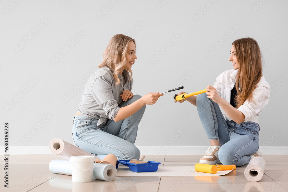 Young women with supplies and wallpaper rolls near grey wall