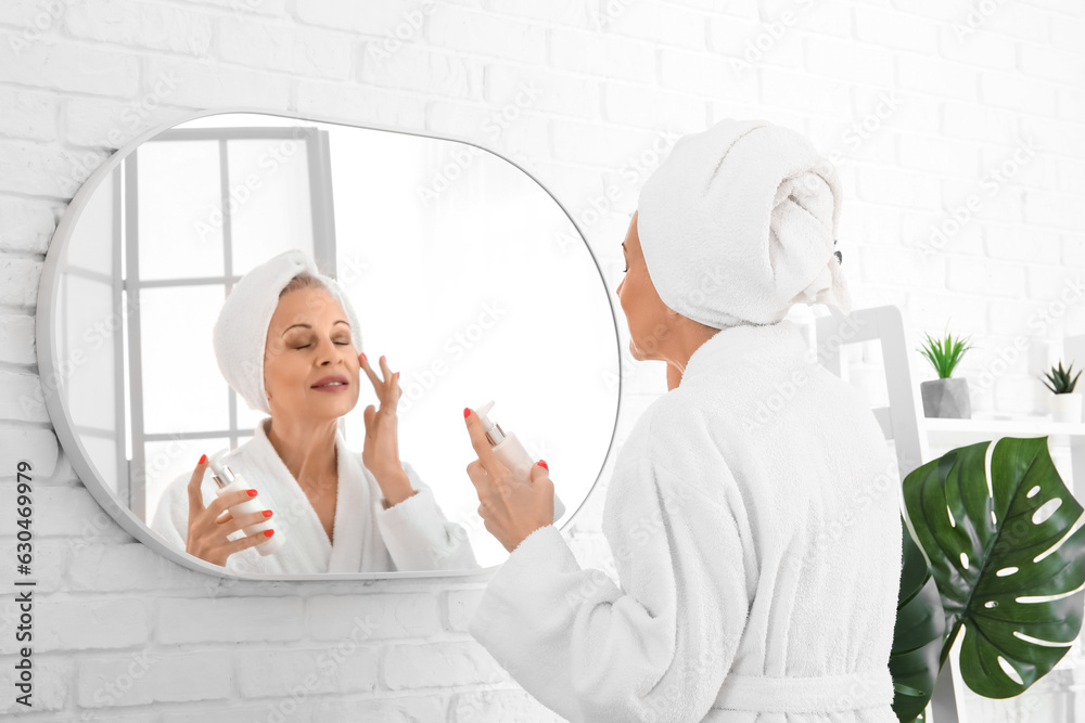 Mature woman applying facial cream near mirror in bathroom