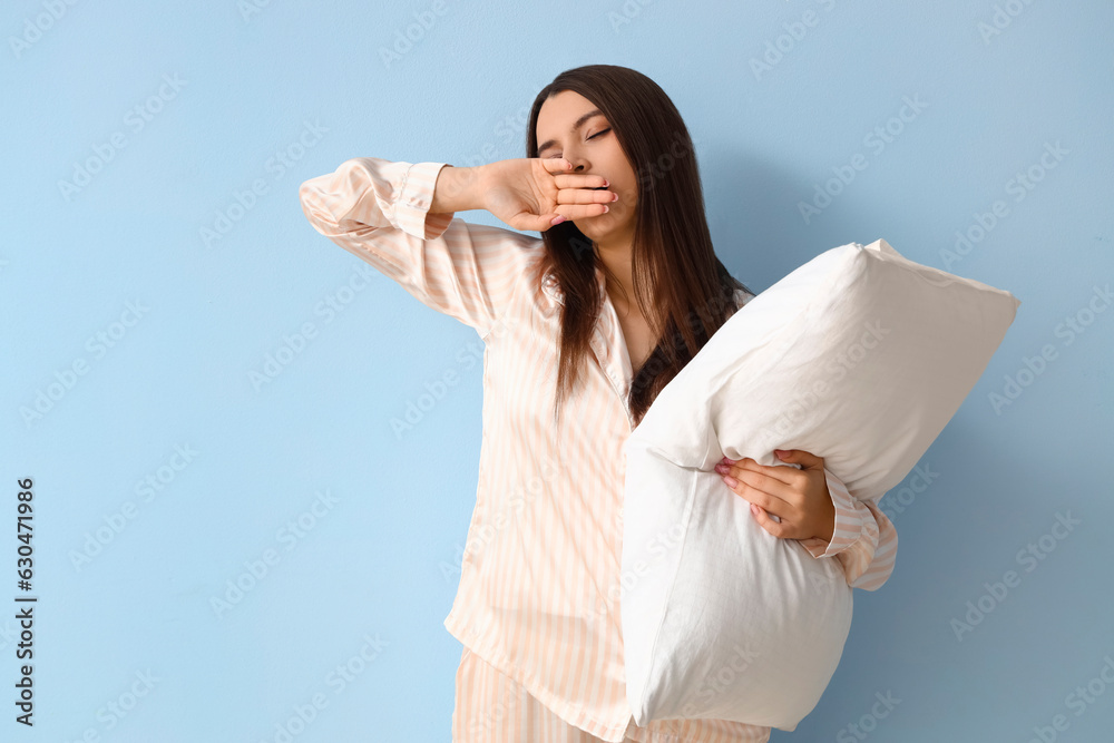 Sleepy young woman in pajamas with pillow on blue background