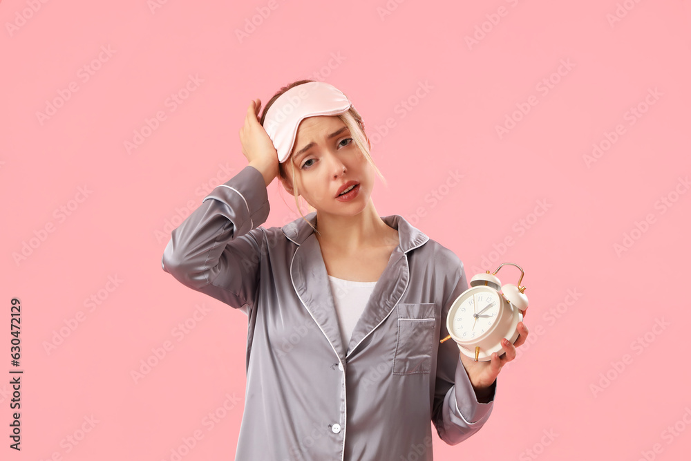 Sleepy young woman in pajamas with alarm clock on pink background