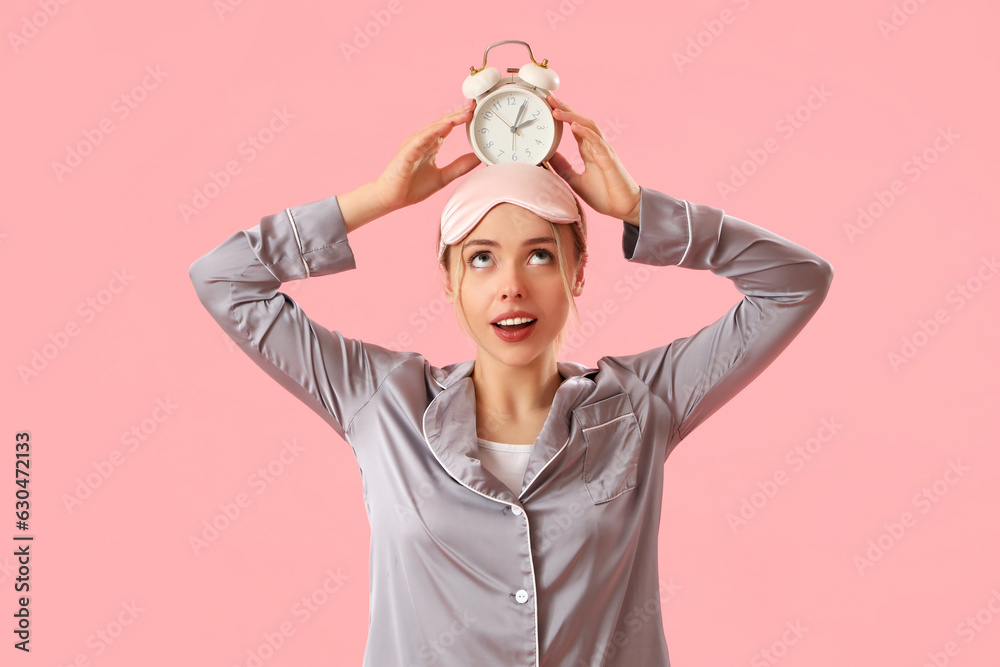 Young woman in pajamas with alarm clock on pink background