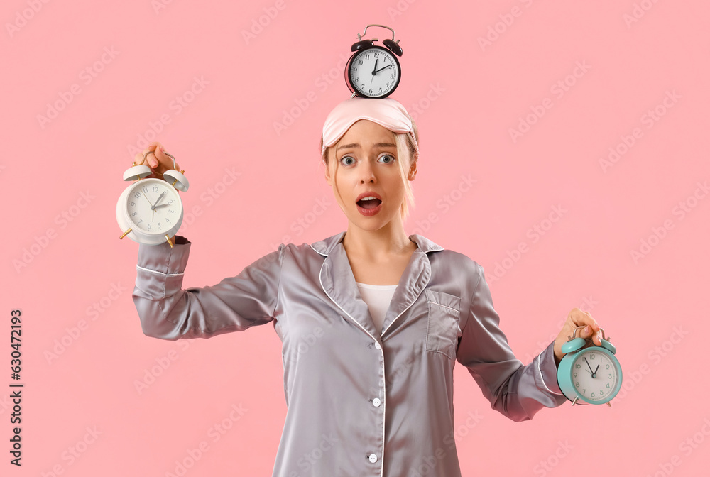 Young woman in pajamas with alarm clocks on pink background