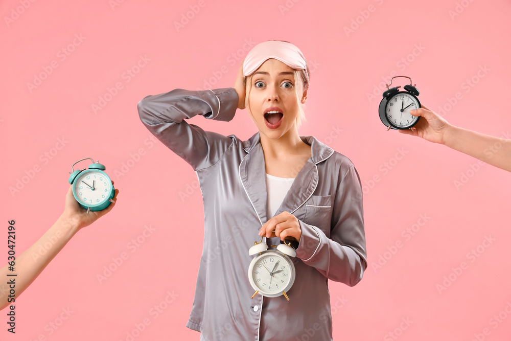 Shocked young woman in pajamas and female hands with alarm clocks on pink background