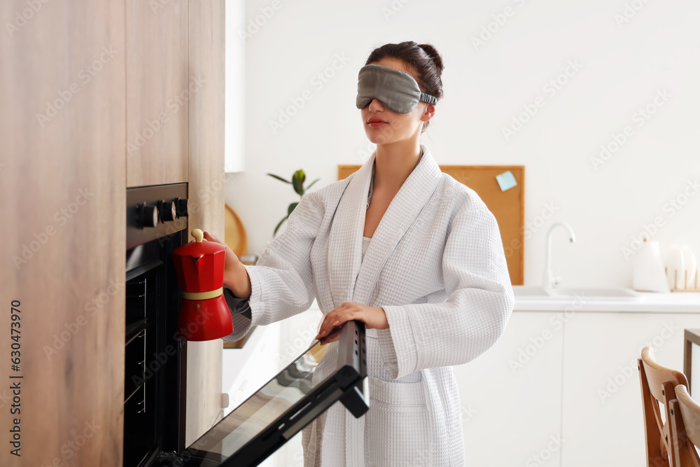 Young woman with sleeping mask putting coffee maker into oven in kitchen