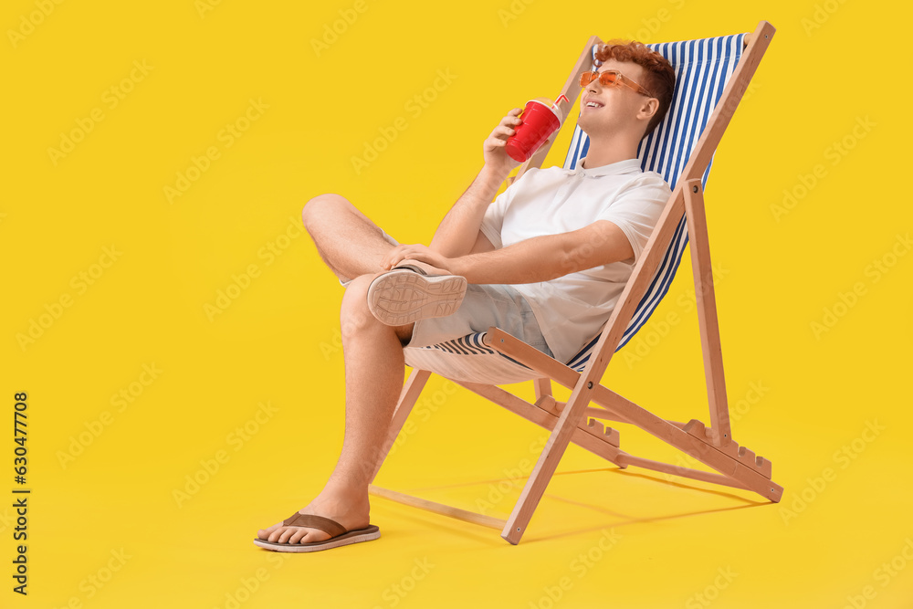 Young man with cup of soda resting in deck chair on yellow background