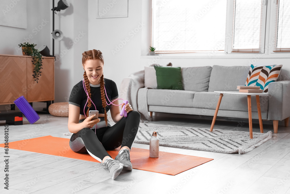 Sporty young woman with skipping rope using mobile phone at home