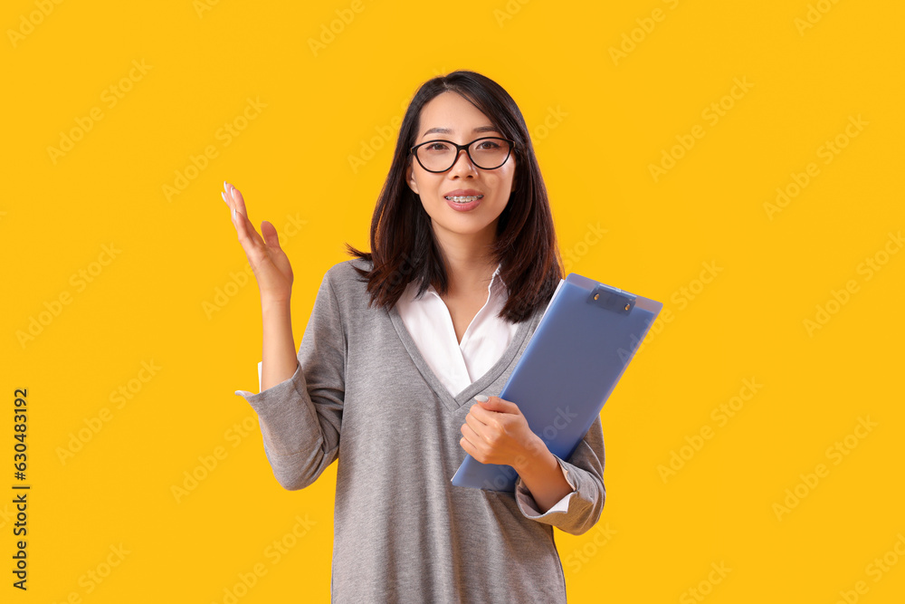 Female Asian teacher with clipboard on yellow background