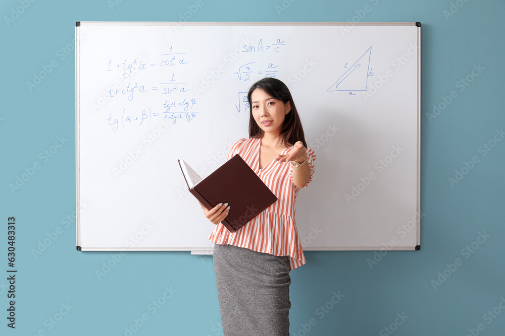 Asian Math teacher with book near flipboard in classroom