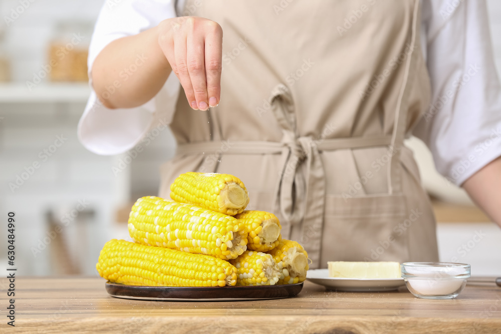 Woman salting boiled corn cobs on wooden table in kitchen