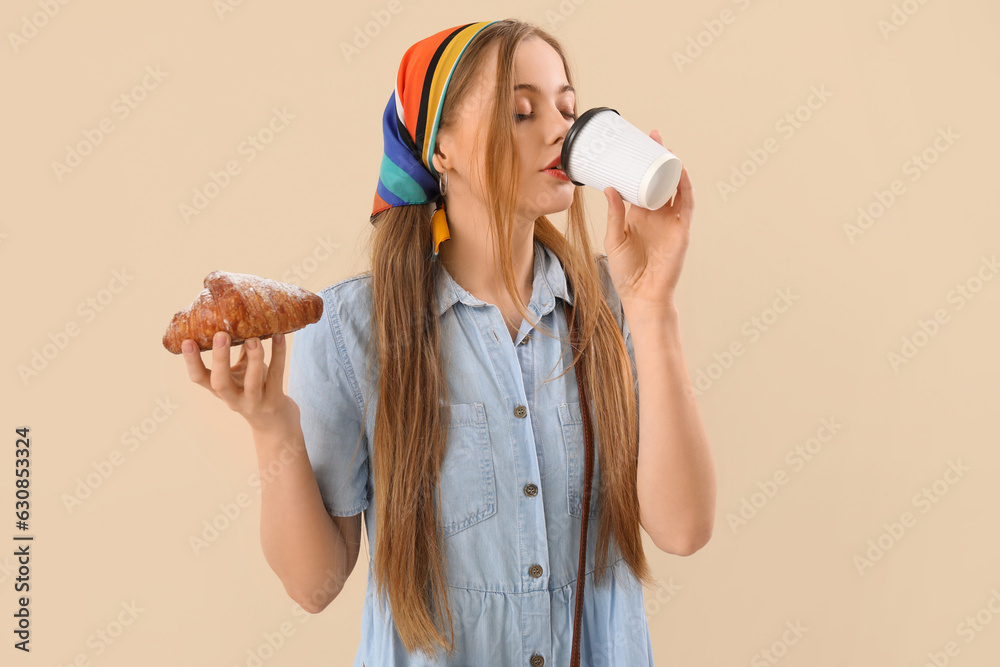 Young woman with tasty croissant drinking coffee on beige background